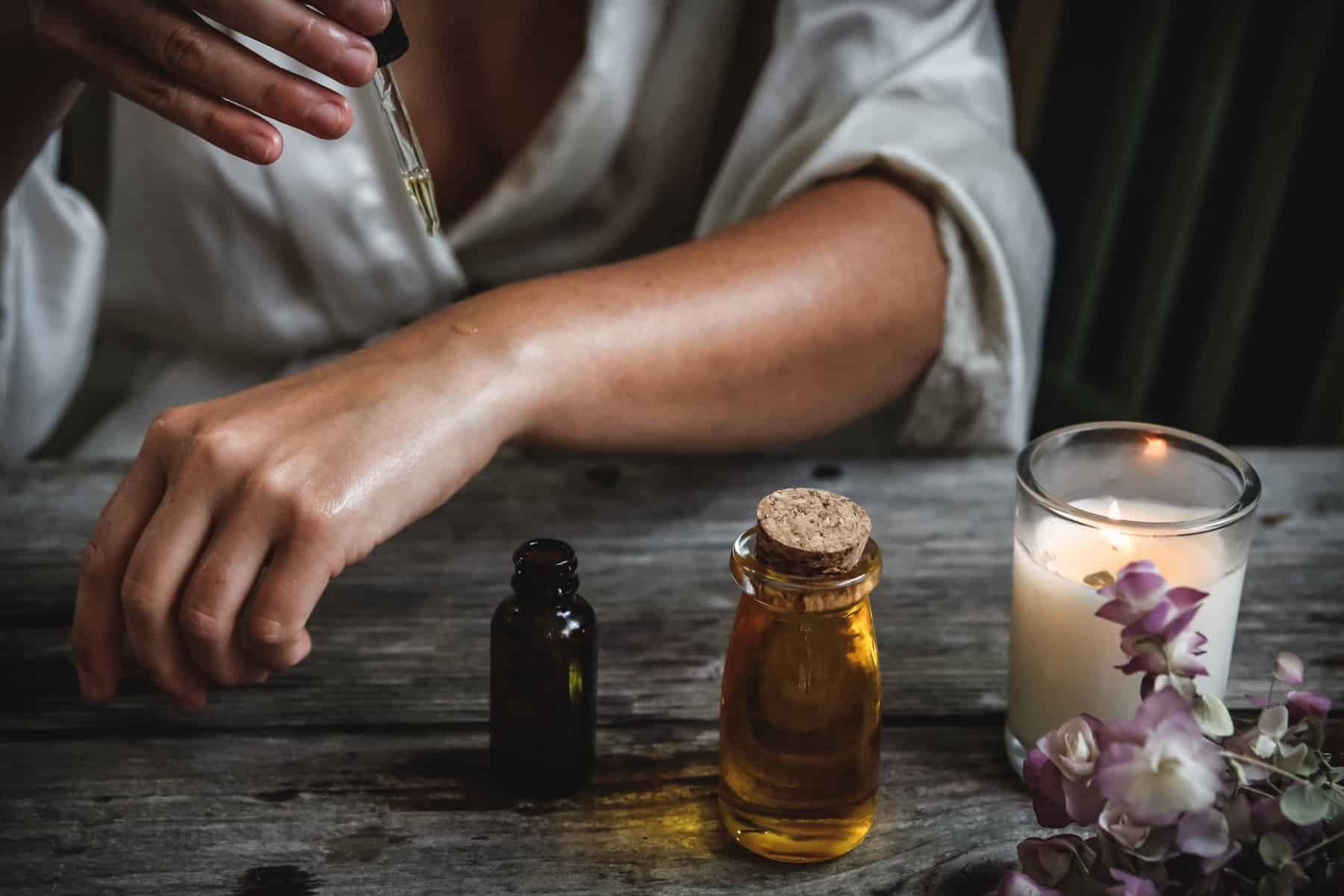 Woman placing oil onto her skin with a dropper with a candle lit next to her